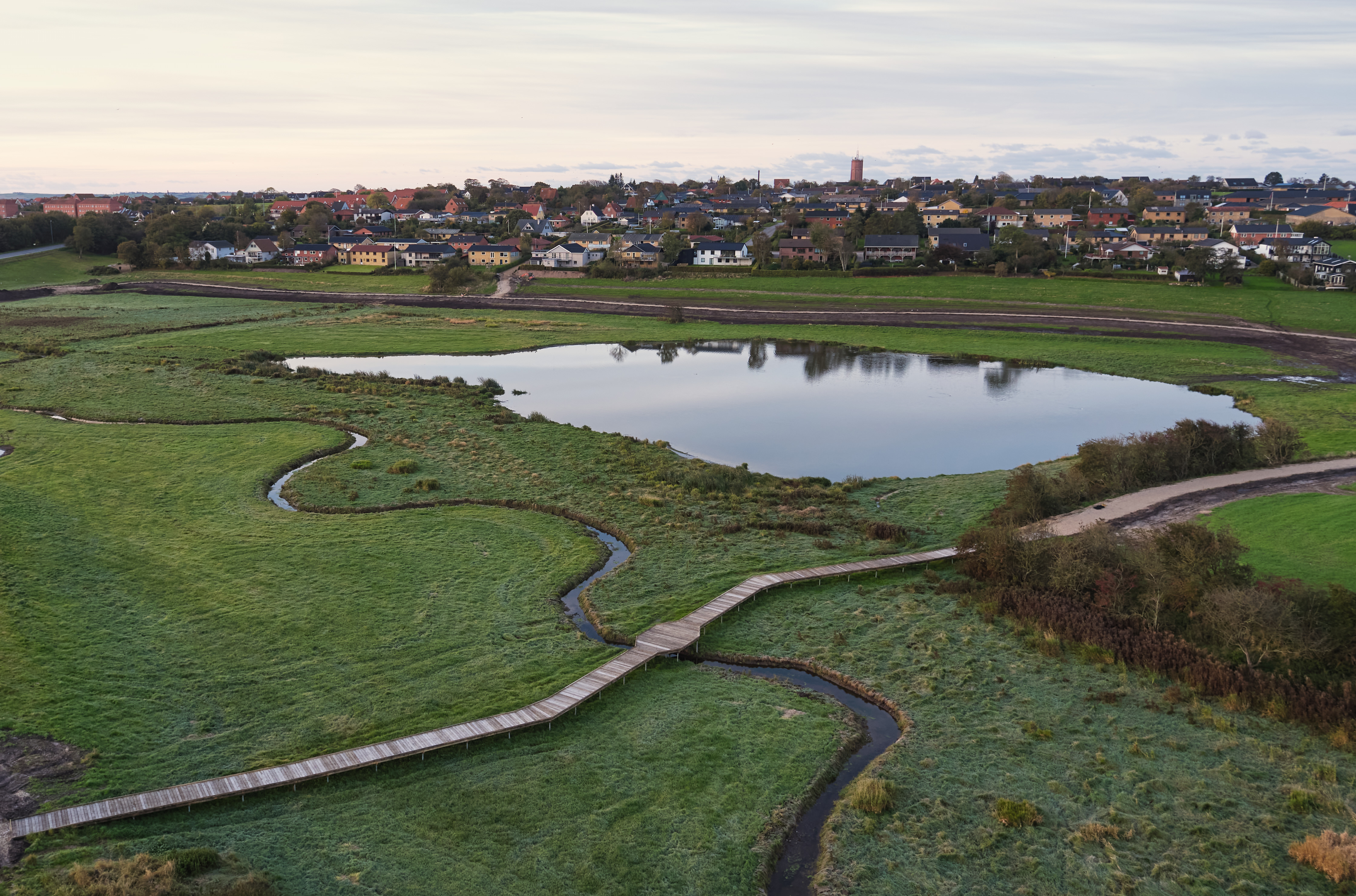 Naturpleieprosjekt i et sumpete område ved en innsjø. En trebro slynger seg gjennom det våte landskapet og gir tilgang uten å forstyrre naturen. I bakgrunnen sees en by med fargerike hus og en kirke.