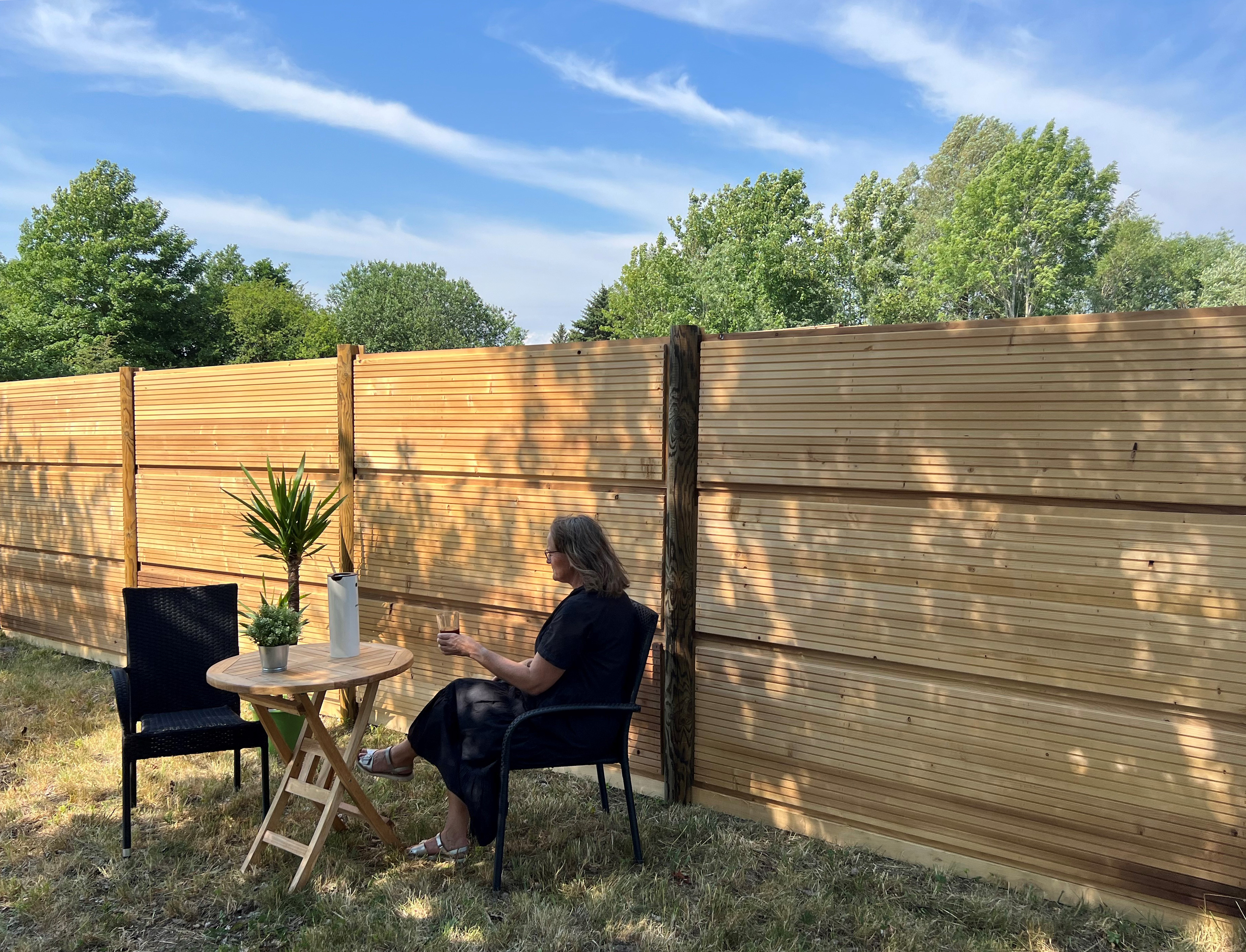 En kvinne sitter ved et bord med en plante og en kanne foran et DeciFence trestøygjerde på en solrik dag.