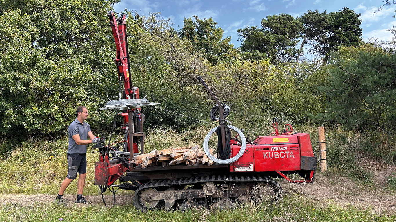 En montør ruller ut tråd til et elektrisk gjerde med en spesialmaskin i et grønt og skogkledd område.