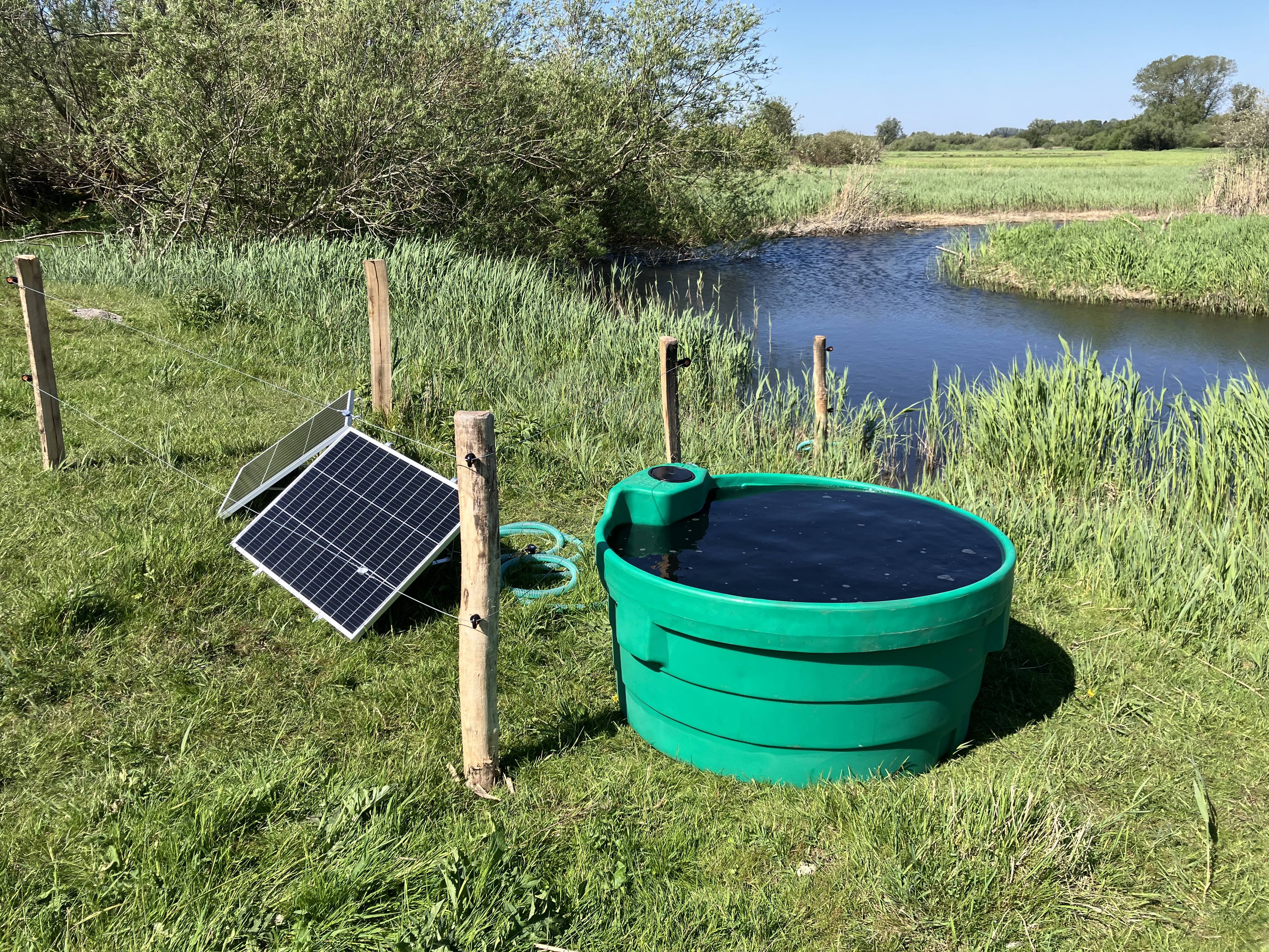 Soldrevet feltpumpe med solcellepanel og vanntank ved en bekk på en eng.