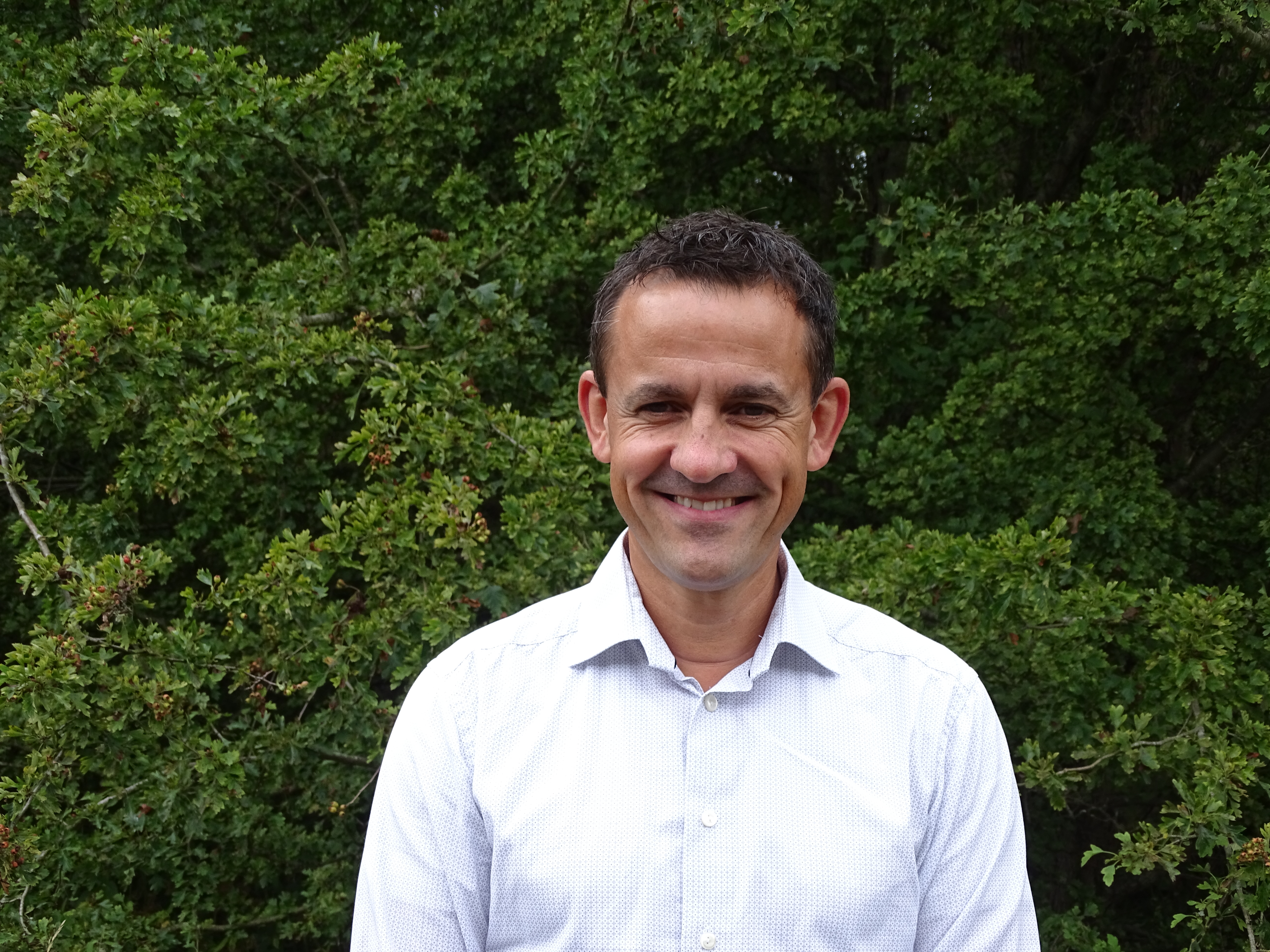 Claus, kontaktperson for Danmark, Sverige og Norge, iført lyseblå skjorte og stående foran grønn vegetasjon.