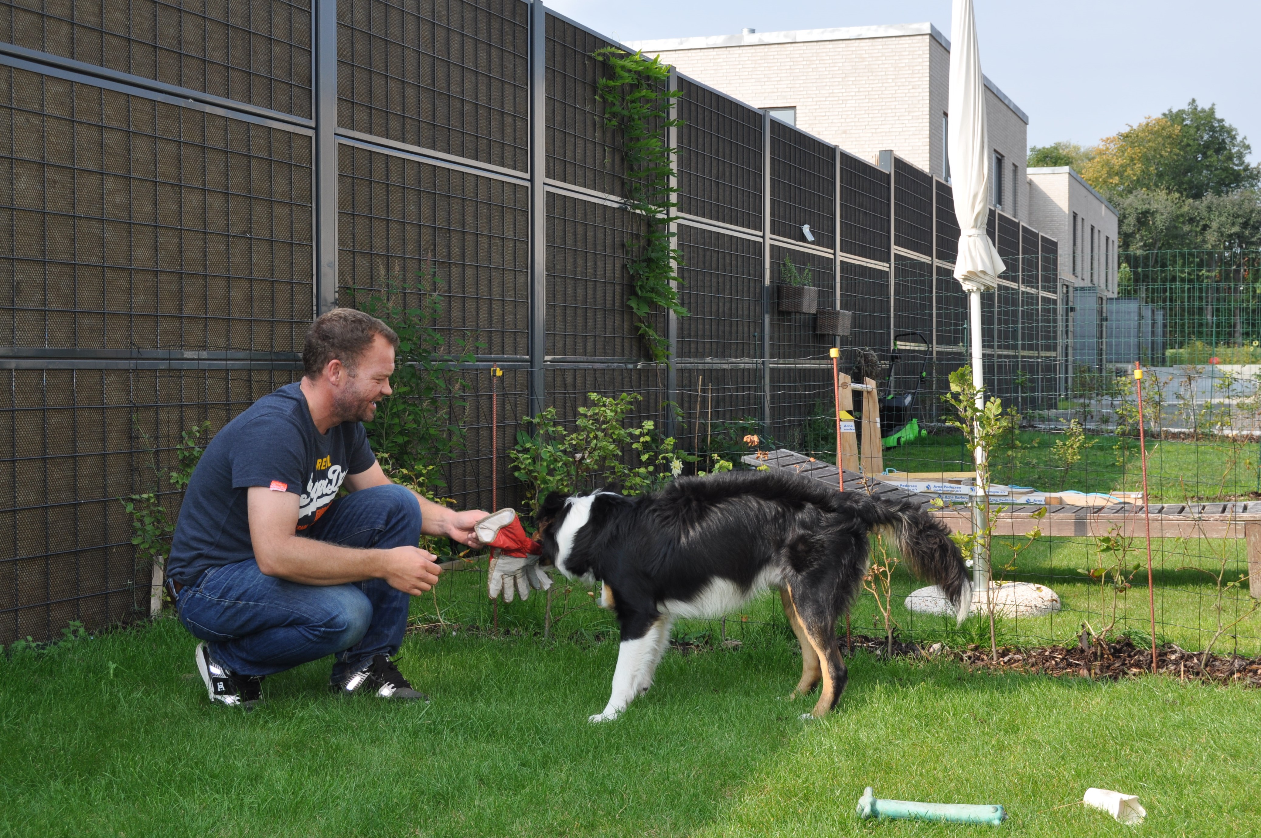 En mann og hunden hans sitter foran et Noistop® Steel-støygjerde fra Poda Gjerder som er installert i en hage for å skjerme for støy fra naboene