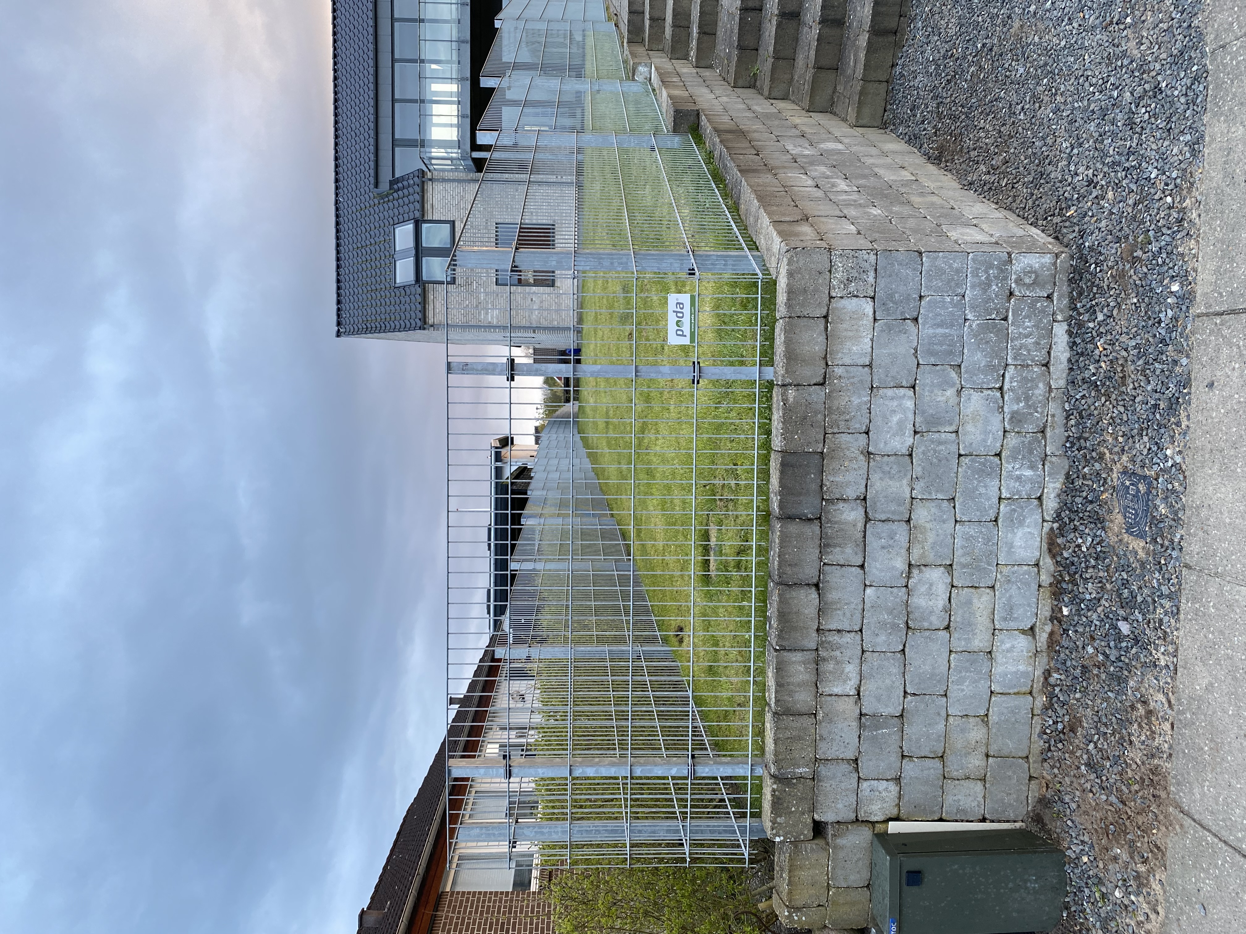 Nærbilde av et panelgjerde montert på en forhøyet plen med en støttemur av betongblokker. I bakgrunnen sees et moderne hus med store glassflater.