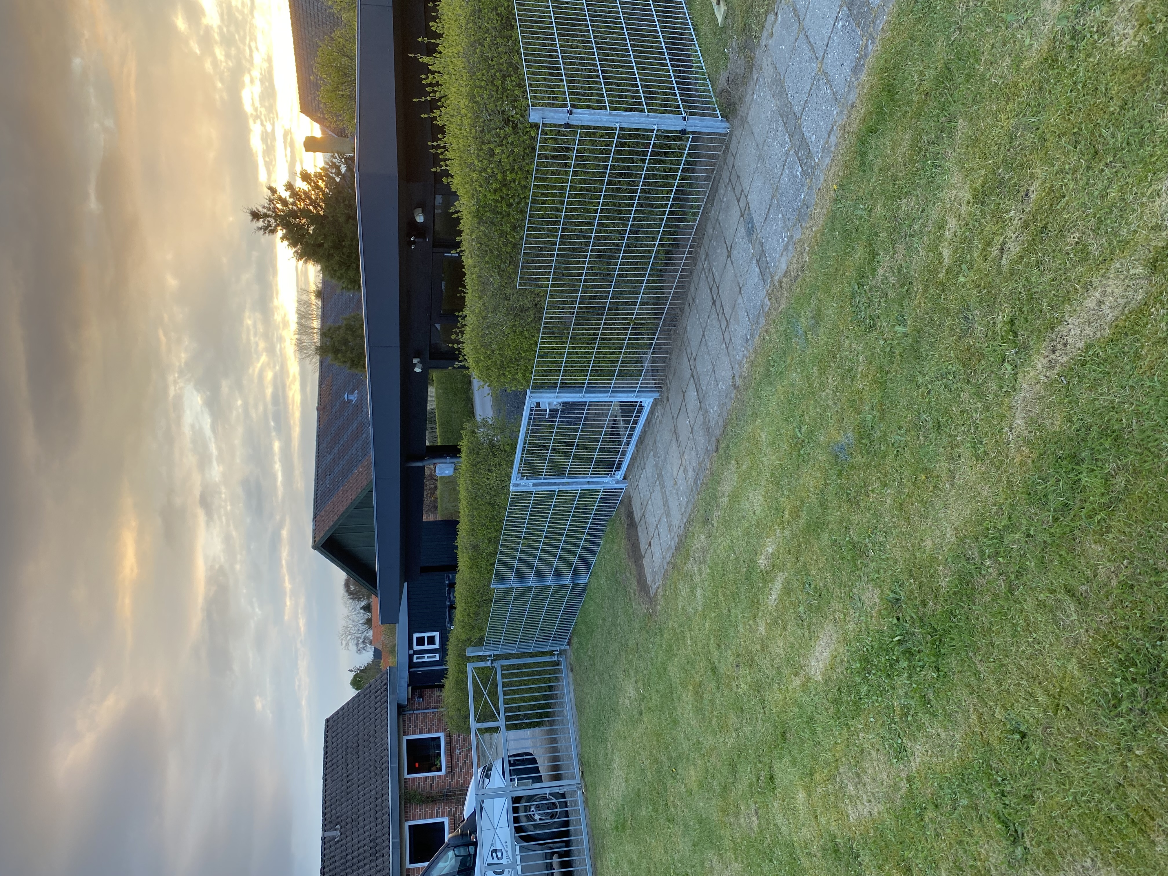Panelgjerde med en port montert langs en sti som skiller en plen fra en hekk. I bakgrunnen sees en carport, flere hus og en solnedgang.